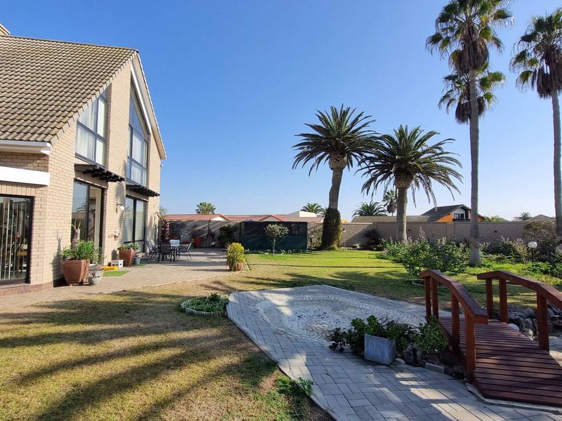 Spacious rear garden featuring a paved patio area, a decorative wooden footbridge, mature palm trees, and a well-maintained brick exterior facade.