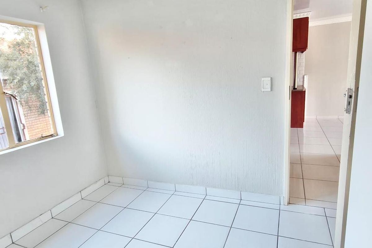 A bright, empty bedroom featuring white tiled flooring, neutral walls, and a view into the adjacent living area.