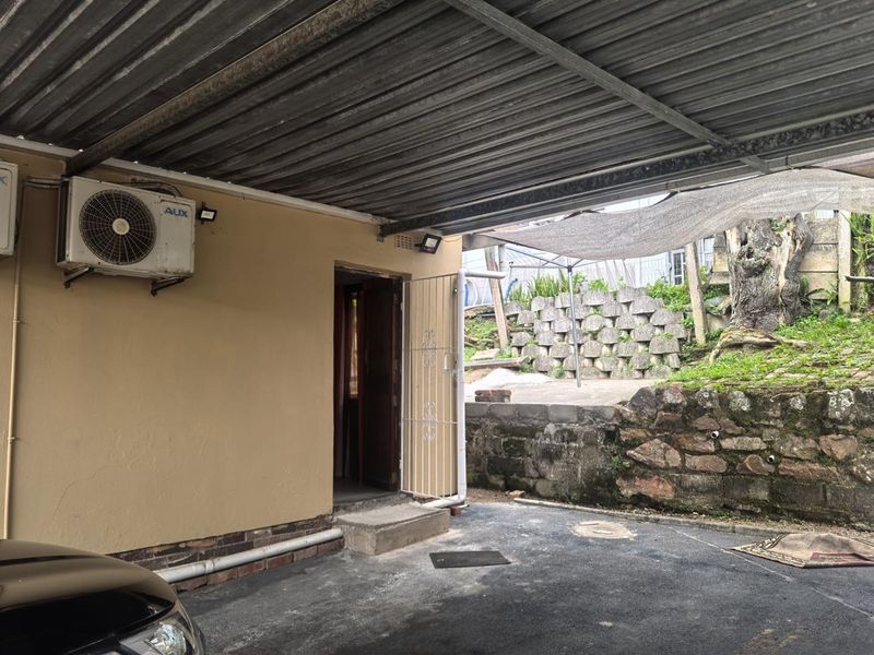 A covered carport area featuring a corrugated metal roof, an exterior air conditioning unit, and a stone retaining wall in the background.
