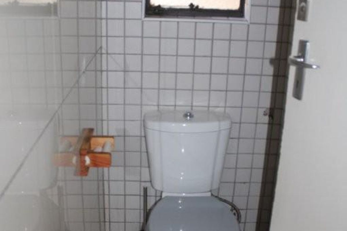 A compact bathroom featuring white wall and floor tiles, a standard toilet, and a small window with security bars.
