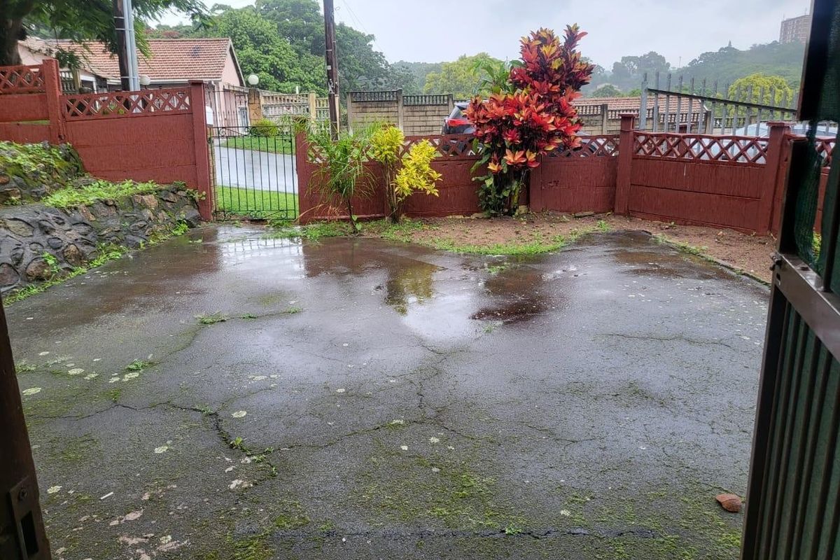 A paved rear courtyard area featuring perimeter fencing, a stone retaining wall, and a metal gate, showing signs of wear and surface water accumulation.