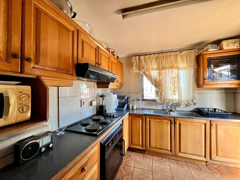 A dated kitchen featuring wooden cabinetry, laminate countertops, an electric hob, and a double bowl stainless steel sink with tiled flooring.