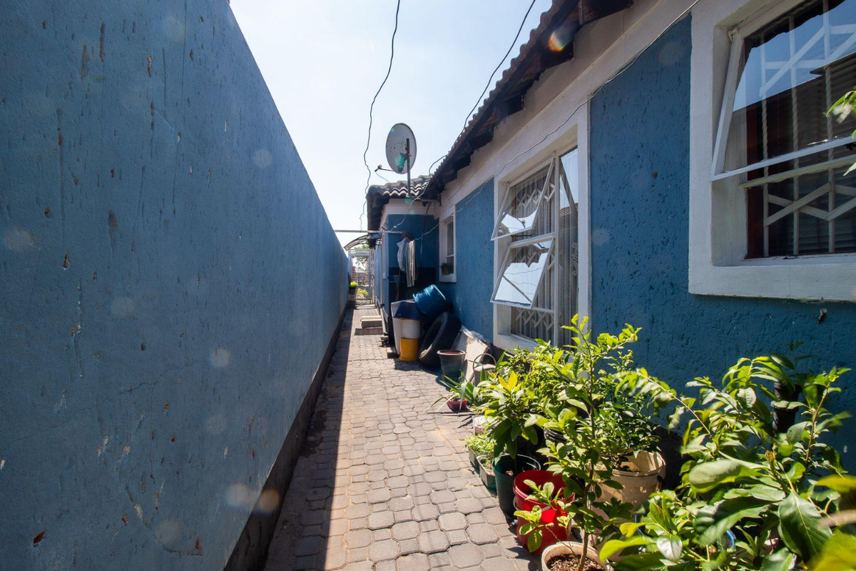 A narrow side passage featuring a paved walkway, blue exterior walls with security bars on windows, and a high boundary wall.