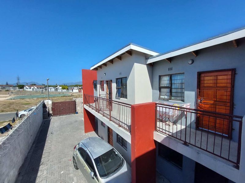 Exterior view of a multi-level apartment building featuring a balcony with metal railings, paved driveway, and perimeter security fencing.