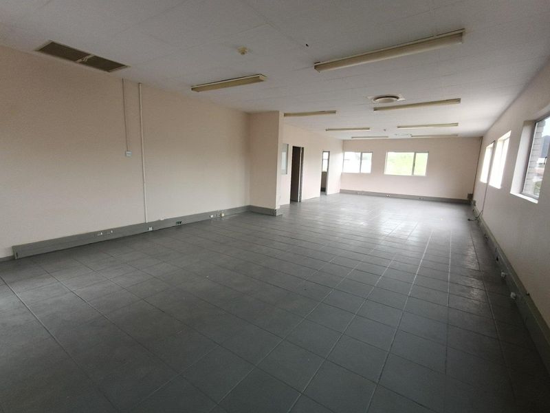 A spacious, empty industrial office area featuring tiled flooring, drop ceilings with integrated lighting, and multiple windows providing natural light.