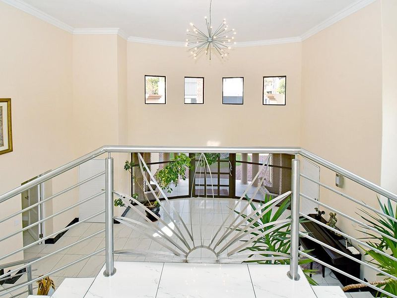 A grand double-volume entrance hall featuring a modern stainless steel balustrade, polished tiled flooring, and a striking feature chandelier.