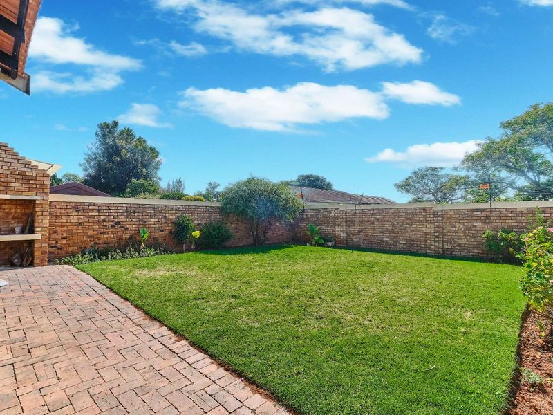 A private rear garden featuring a well-maintained lawn, paved patio area, and a built-in brick braai, enclosed by a secure perimeter wall.