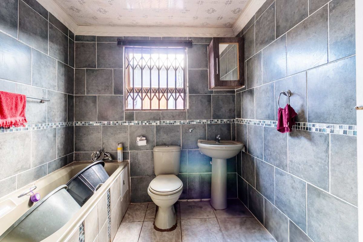 A functional bathroom featuring full-height grey wall tiling, a pedestal basin, a bathtub, and a window with security bars.