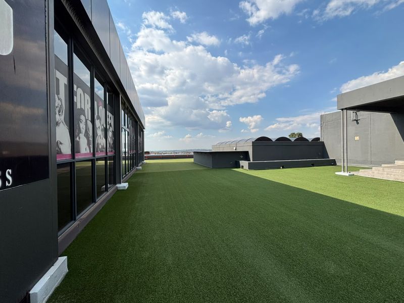A spacious rooftop terrace featuring artificial turf, modern metal cladding, and extensive glass paneling, offering an open-air environment suitable for office breakout space.