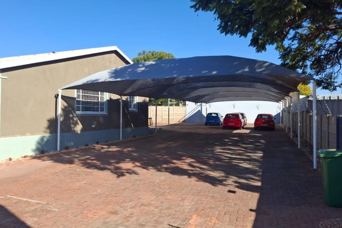 Rear exterior view of the office property featuring a large shade cloth carport structure over a paved parking area and secure perimeter fencing.