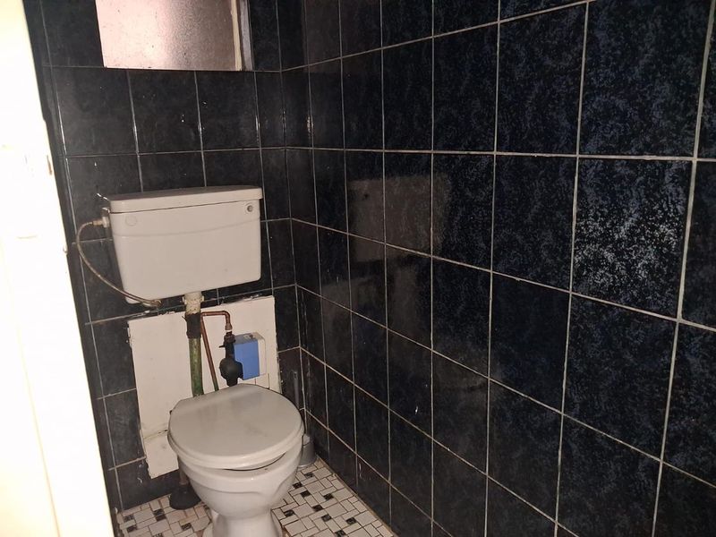 A small, dated bathroom featuring dark wall tiles, mosaic floor tiling, and a standard toilet with exposed plumbing.