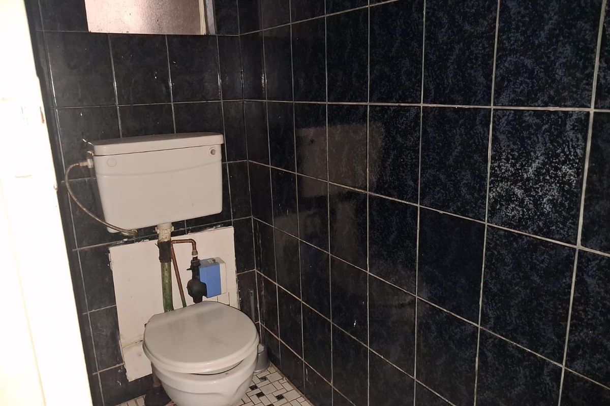 A small, dated bathroom featuring dark wall tiles, mosaic floor tiling, and a standard toilet with exposed plumbing.