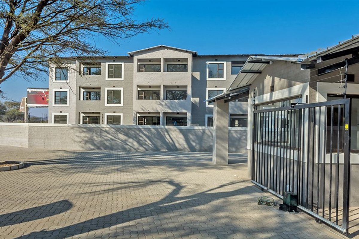 A view of the apartment complex entrance featuring a paved driveway, secure gated access, and perimeter walling with electric fencing.