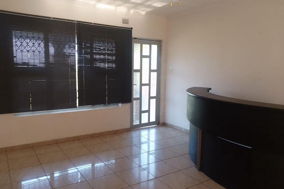 A bright, tiled room featuring a built-in reception-style counter, large windows with blinds, and a glass-paneled exterior door.
