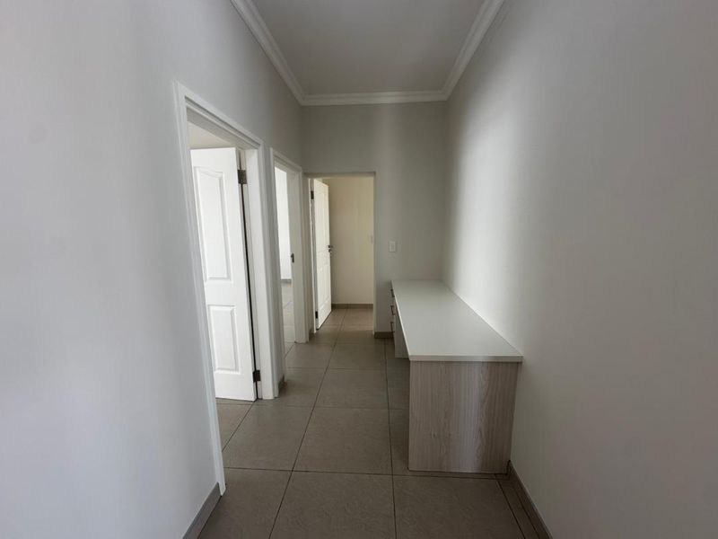A clean, neutral hallway featuring tiled flooring, cove cornicing, and a built-in desk area, providing a functional transition space between rooms.