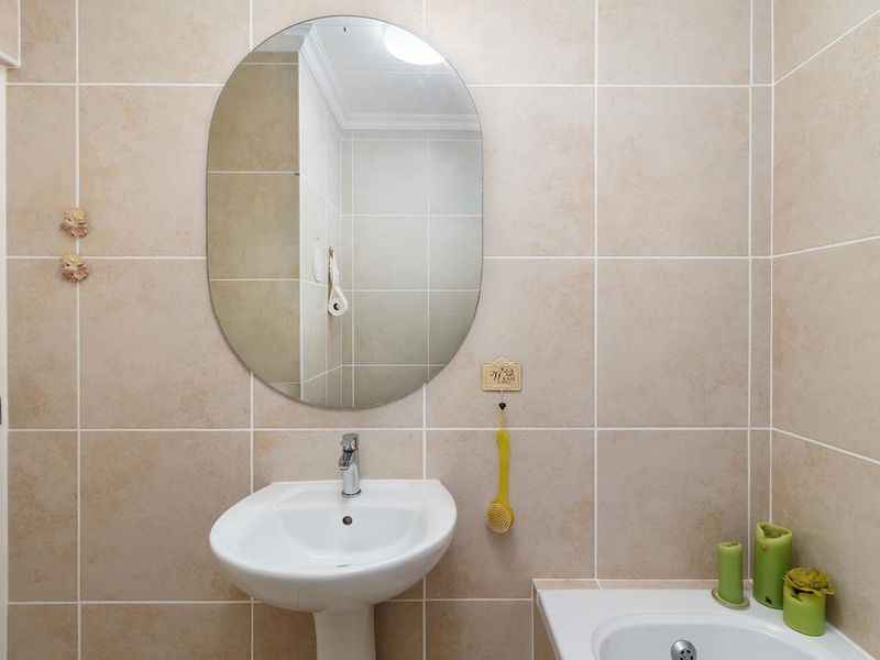 A clean and functional bathroom featuring a pedestal basin, chrome mixer tap, and neutral ceramic wall tiling with an oval mirror.