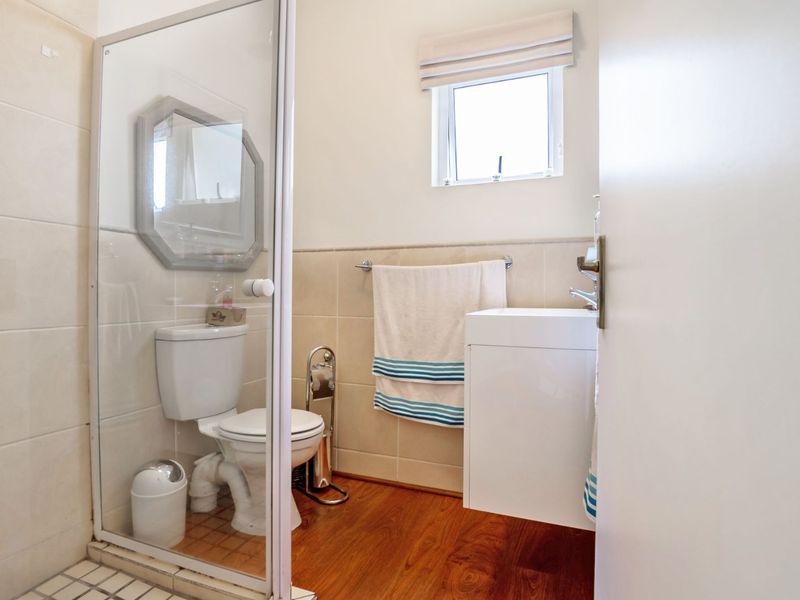 A compact bathroom featuring a glass-enclosed shower, toilet, basin, and neutral wall tiling with warm timber flooring.