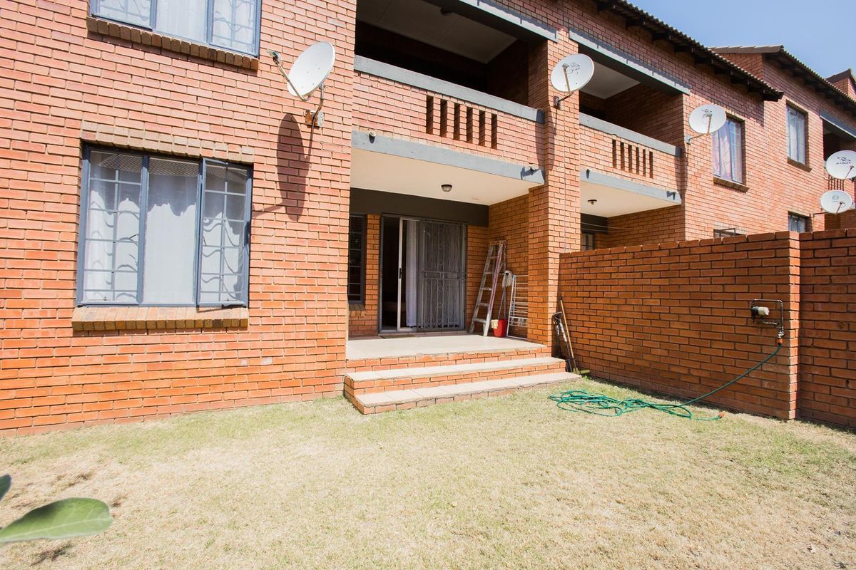 A rear view of a brick townhouse featuring a covered patio, small private garden, and upper-level balcony with security gates.