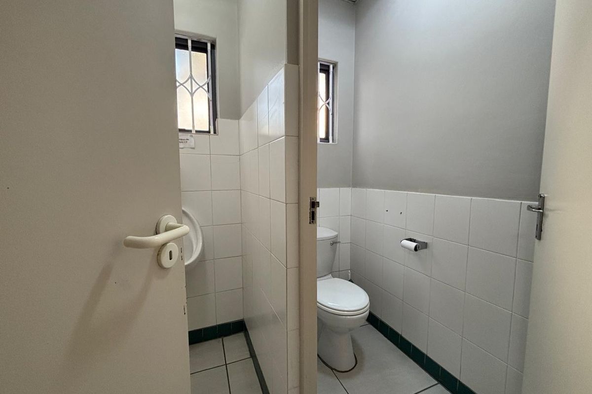 A functional office restroom featuring a separate urinal and toilet stall with tiled finishes and security bars on the windows.