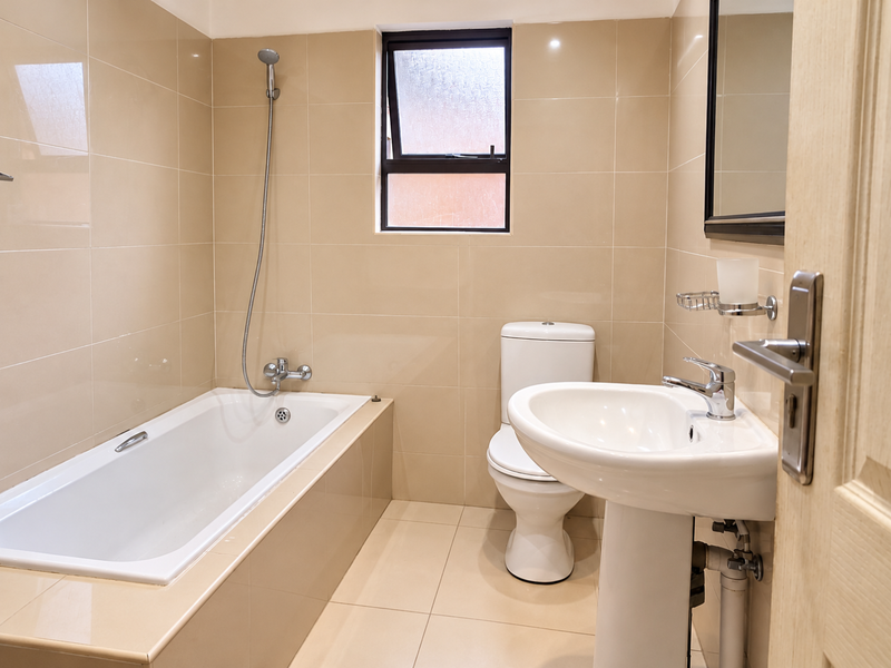 A clean, functional bathroom featuring a bathtub with handheld shower, pedestal sink, and toilet, finished with neutral wall and floor tiles.
