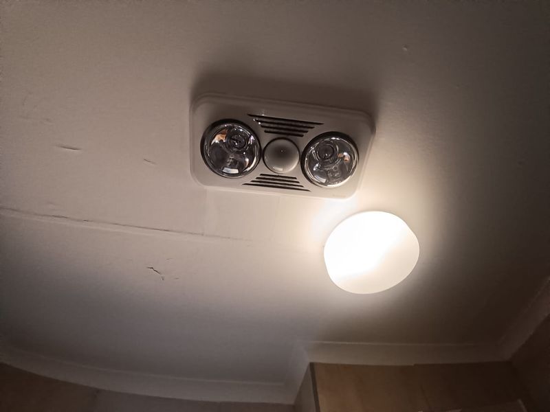 A close-up view of a bathroom ceiling featuring a heat lamp and exhaust fan unit, showing visible cracks and peeling paint in the plaster.