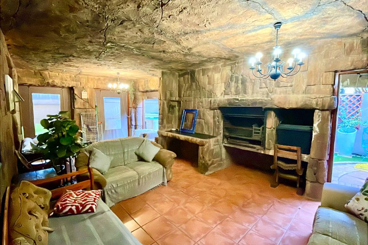 A rustic living area featuring unique stone-clad walls, a textured stone ceiling, tiled flooring, and a built-in stone fireplace with a wrought iron chandelier.