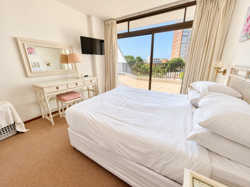 A bright bedroom featuring carpet flooring, a wall-mounted television, and large sliding glass doors that open onto a private balcony.