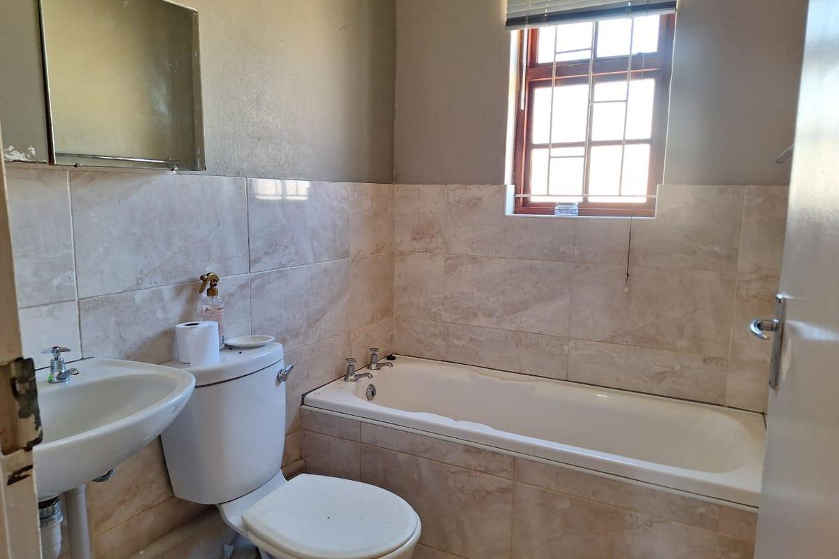 A functional bathroom featuring a standard bathtub, pedestal basin, and toilet with neutral wall tiling and a window with security bars.