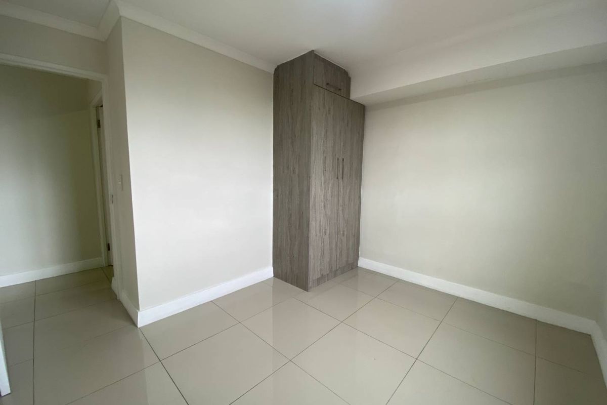 A clean, empty bedroom featuring neutral tiled flooring, white walls, and a built-in wooden wardrobe with overhead storage.