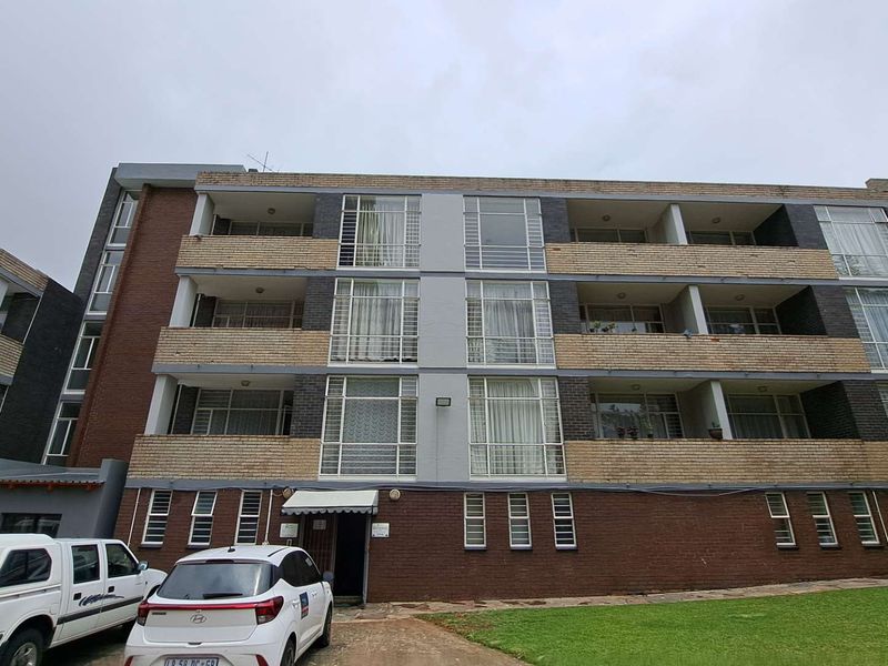 A multi-story apartment building featuring a brick facade, integrated balconies, and large steel-framed windows, set against a landscaped lawn area.