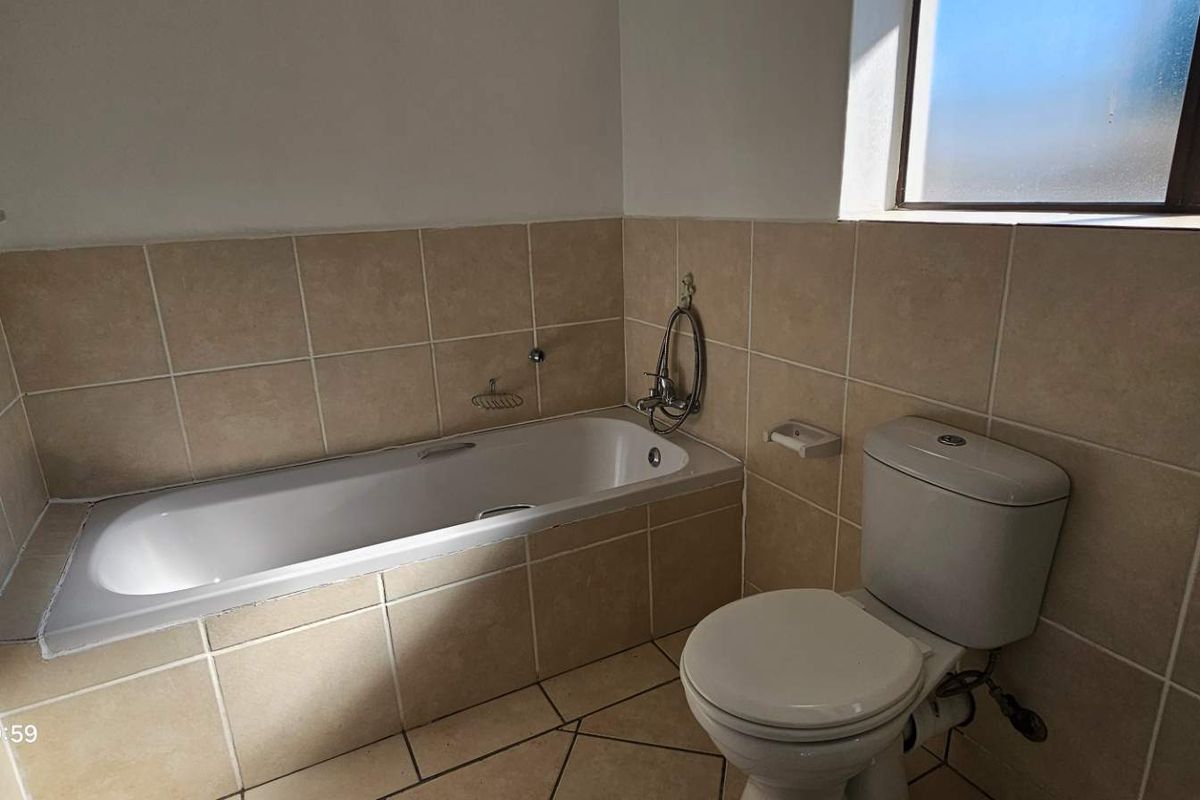A functional bathroom featuring a built-in bathtub, toilet, and neutral ceramic wall and floor tiles, offering a clean and practical space.