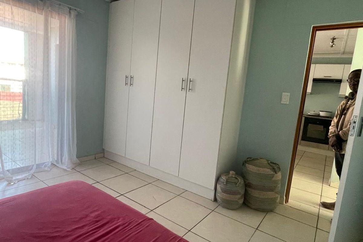 A bedroom featuring built-in white cupboards and tiled flooring, with a view through the doorway into the kitchen area.