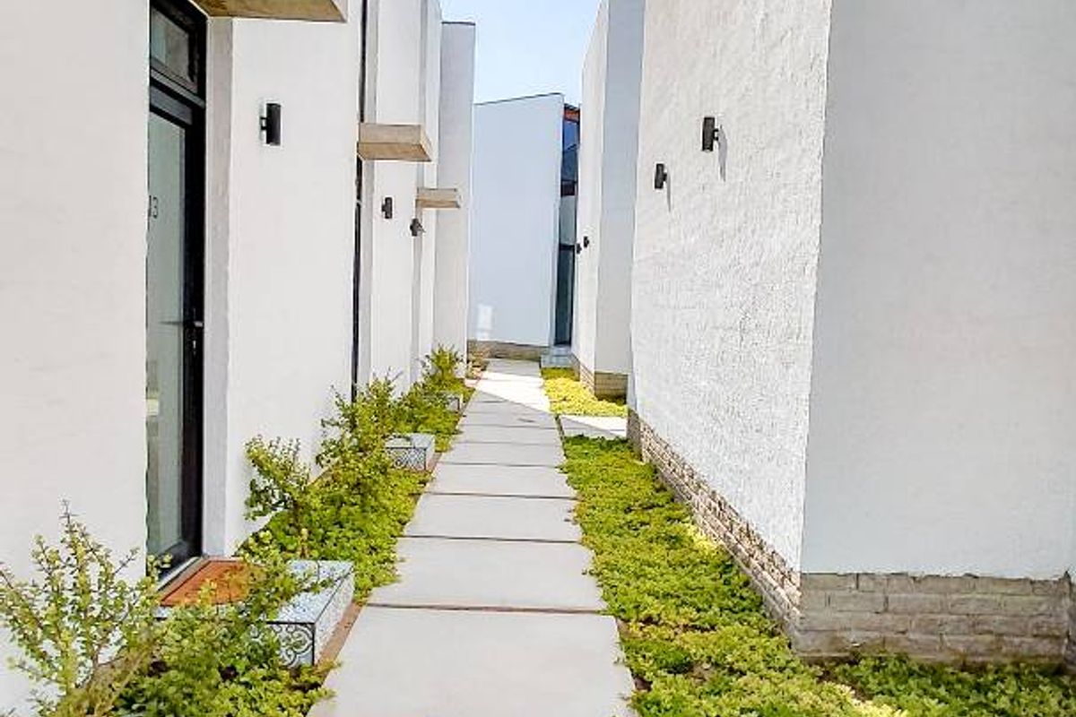 A modern exterior walkway featuring concrete pavers, landscaped garden beds, and contemporary wall-mounted lighting fixtures between apartment units.