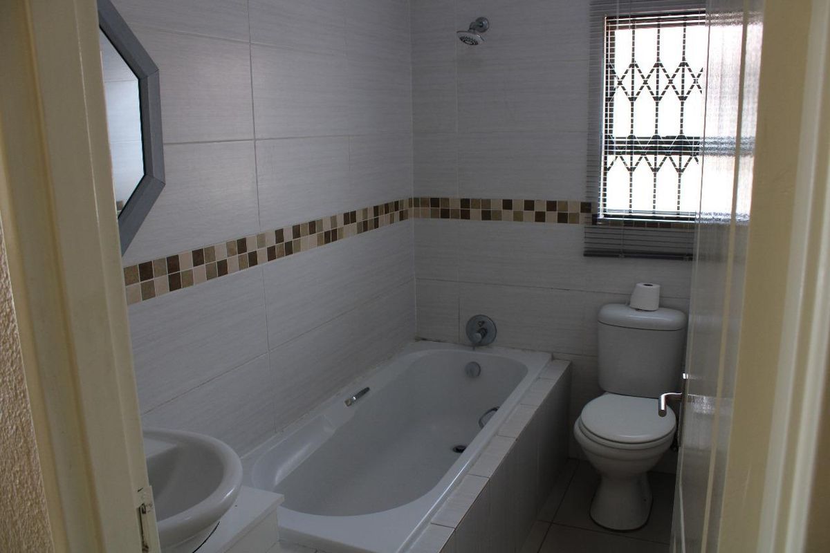 A functional bathroom featuring a built-in bathtub, wall-mounted shower head, pedestal basin, and a toilet with a window featuring security bars.