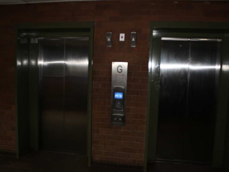 A dimly lit apartment building hallway featuring two stainless steel elevator doors set into an exposed brick wall.