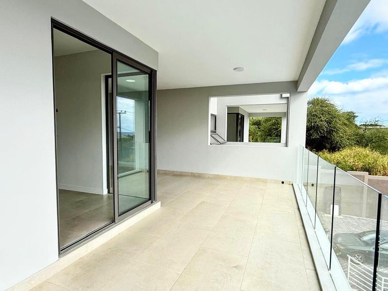 A spacious, modern covered balcony featuring large-format floor tiles, a sleek frameless glass balustrade, and sliding door access to the interior living space.