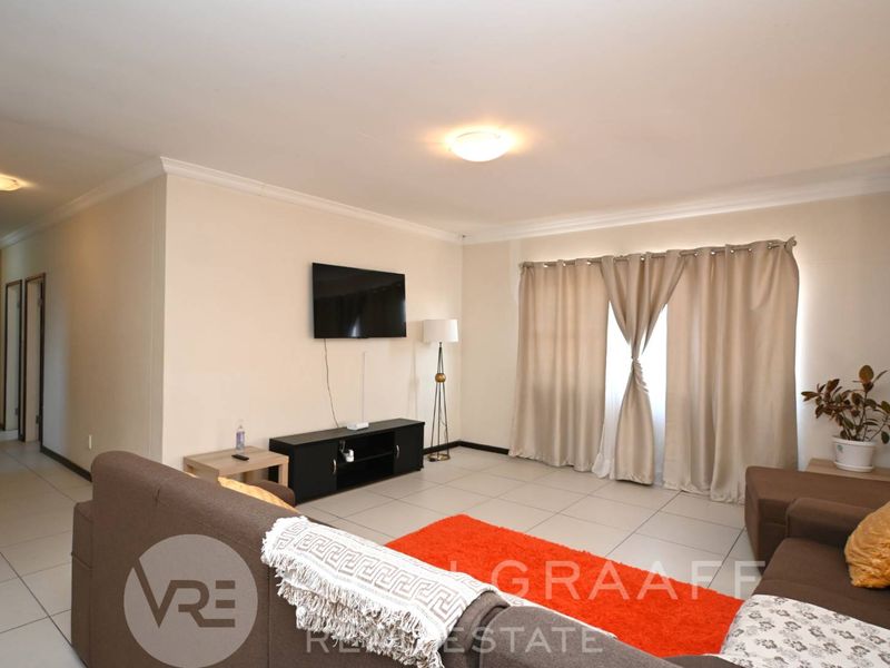 A spacious living room featuring neutral tiled flooring, cove cornicing, and ample natural light through large window treatments.