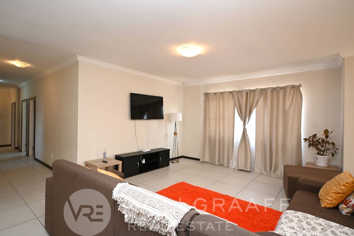 A spacious living room featuring neutral tiled flooring, cove cornicing, and ample natural light through large window treatments.
