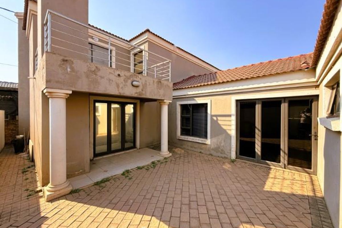 Rear exterior view of the house featuring a paved courtyard, a covered patio area with columns, and an upper-level balcony with metal railings.