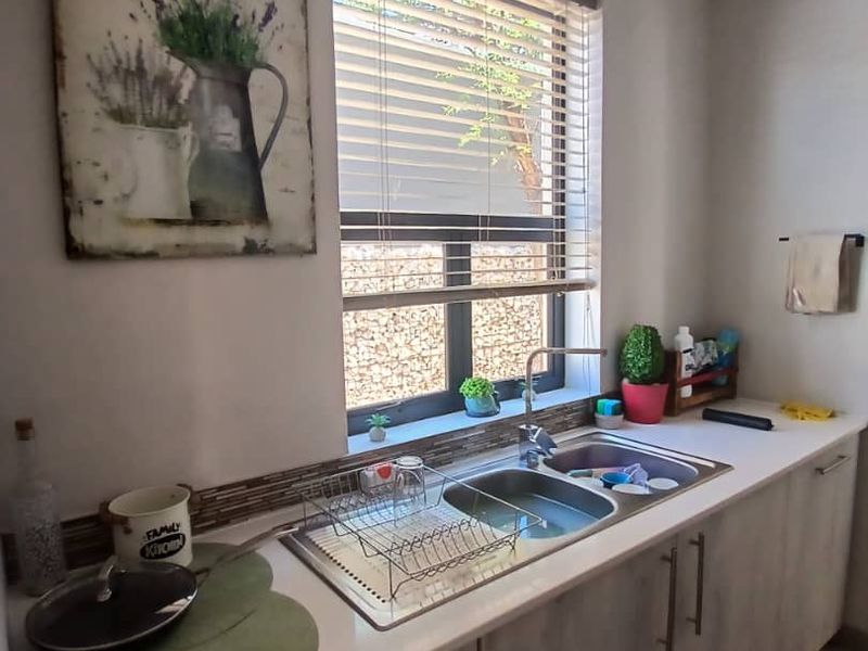 A functional kitchen area featuring a double bowl stainless steel sink, modern laminate cabinetry, and a tiled splashback under a window with venetian blinds.