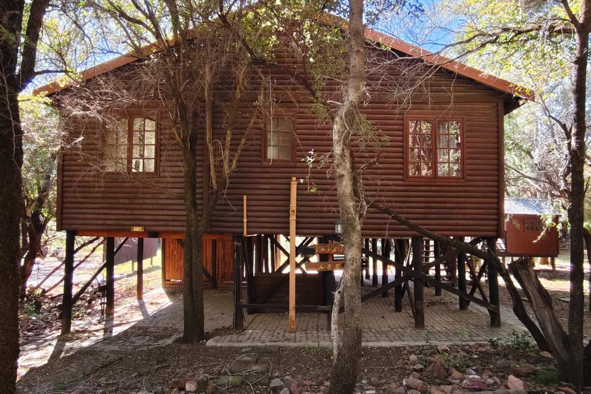 A rustic elevated log cabin style house featuring timber wall construction, raised support pillars, and a paved ground level area surrounded by mature trees.