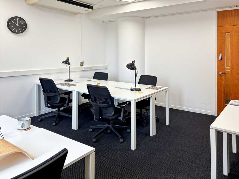 A functional office space featuring dark carpet flooring, a suspended ceiling with integrated air conditioning, and a solid wooden door.