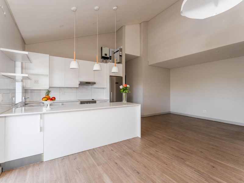 Modern open-plan kitchen and living area featuring sleek white cabinetry, stone countertops, integrated appliances, and light wood-look laminate flooring.