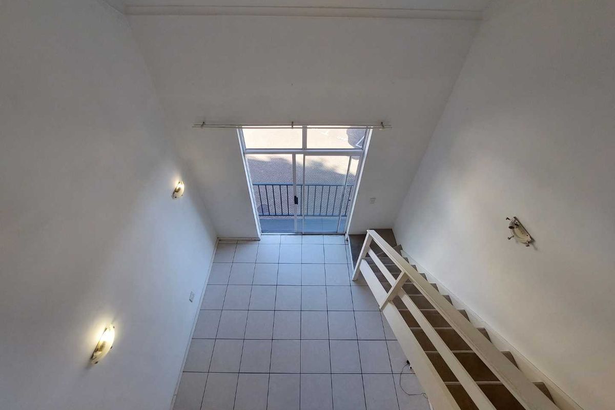 An empty, high-ceiling living area featuring tiled flooring, a staircase with a balustrade, and a sliding glass door leading to a balcony.