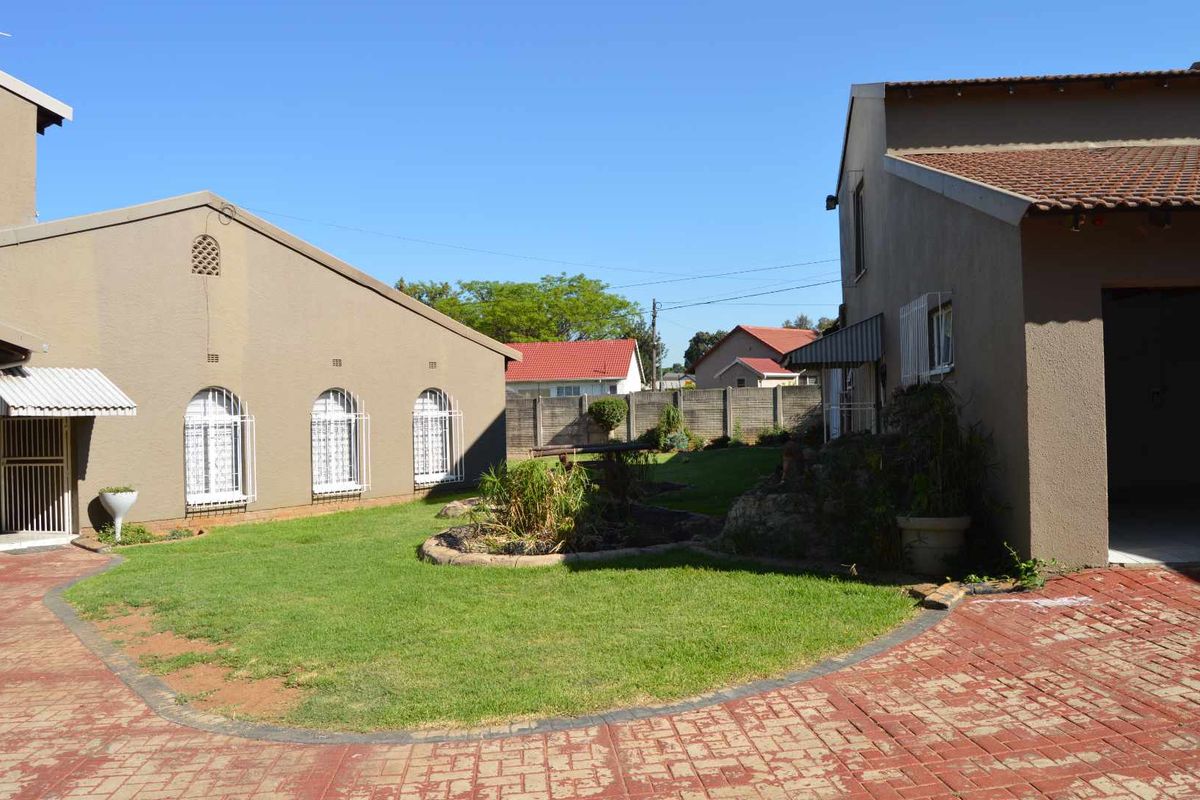 A view of the rear garden area featuring a paved driveway, well-maintained lawn, and secure building facades with tiled roofing.