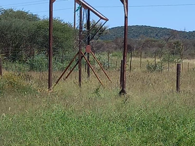 An outdoor view of a weathered metal gantry structure situated in a rural field with overhead power lines and wire fencing.