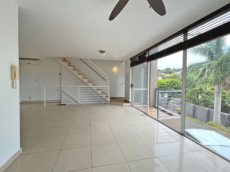 Spacious open-plan living area featuring large sliding glass doors leading to a balcony, tiled flooring, a staircase, and a wall-mounted air conditioning unit.