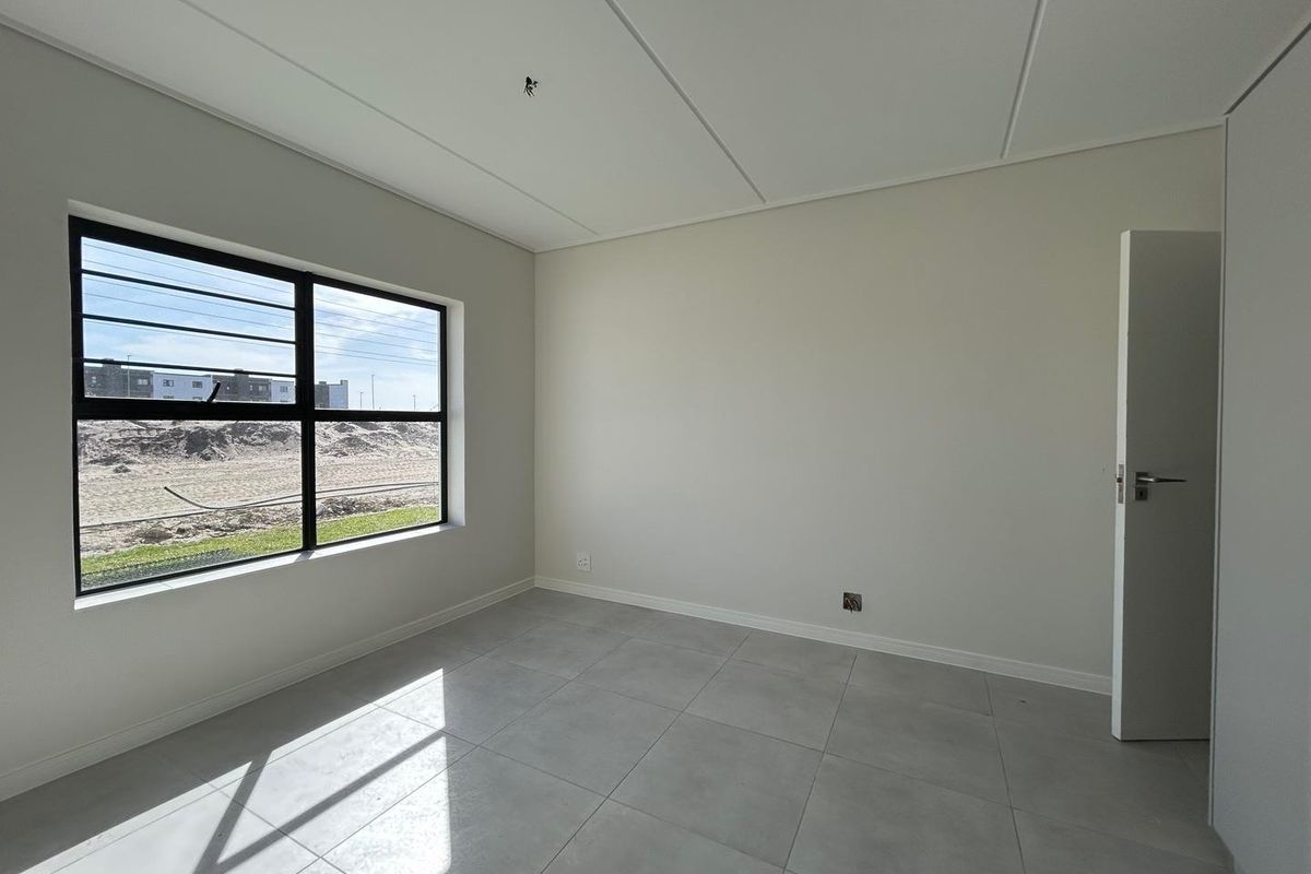 A bright, empty bedroom featuring modern grey floor tiles, a large window with black frames, and a clean, neutral interior finish.