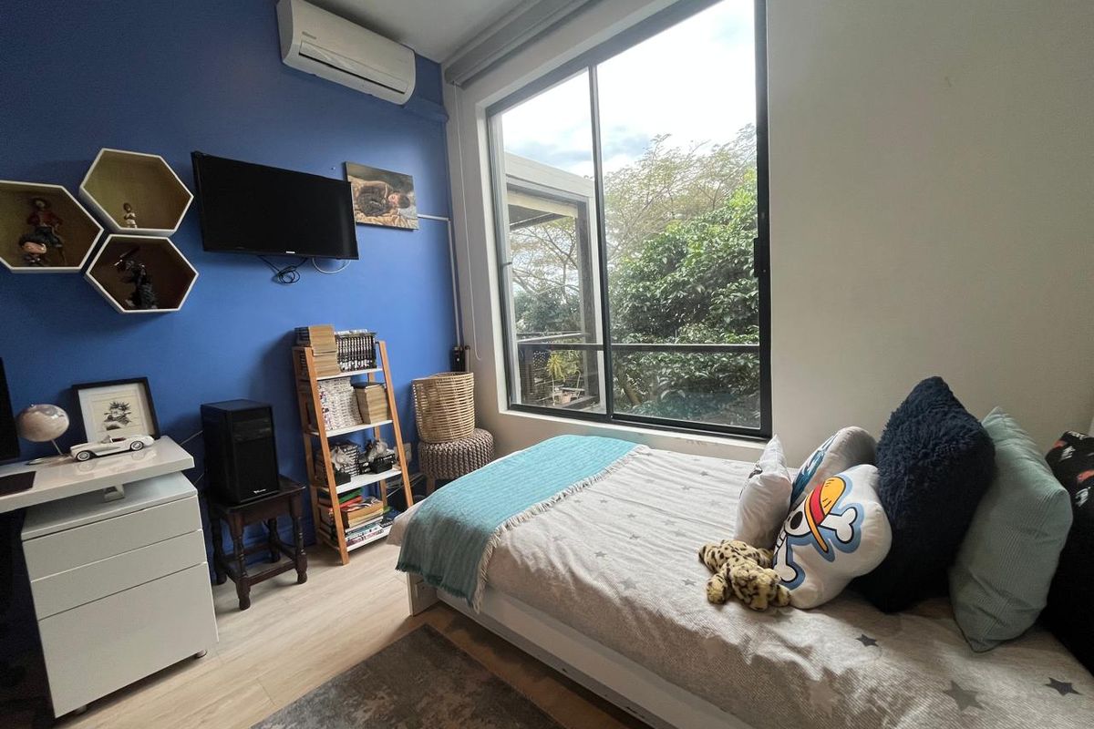 A well-lit bedroom featuring a large window with garden views, light wood-look flooring, and a wall-mounted air conditioning unit.
