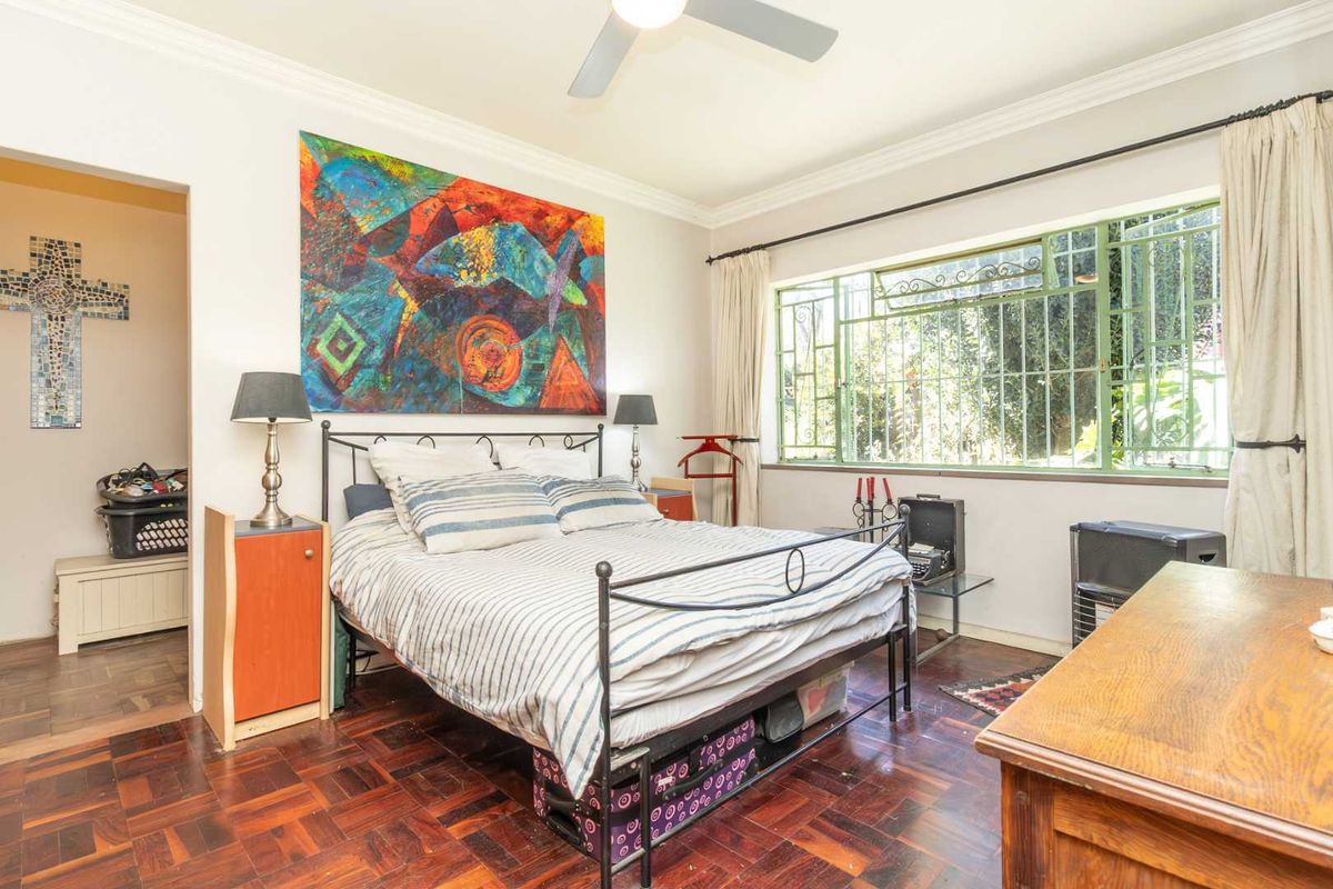 A bedroom featuring parquet flooring, a ceiling fan, and large windows with security bars, offering a view of the garden.